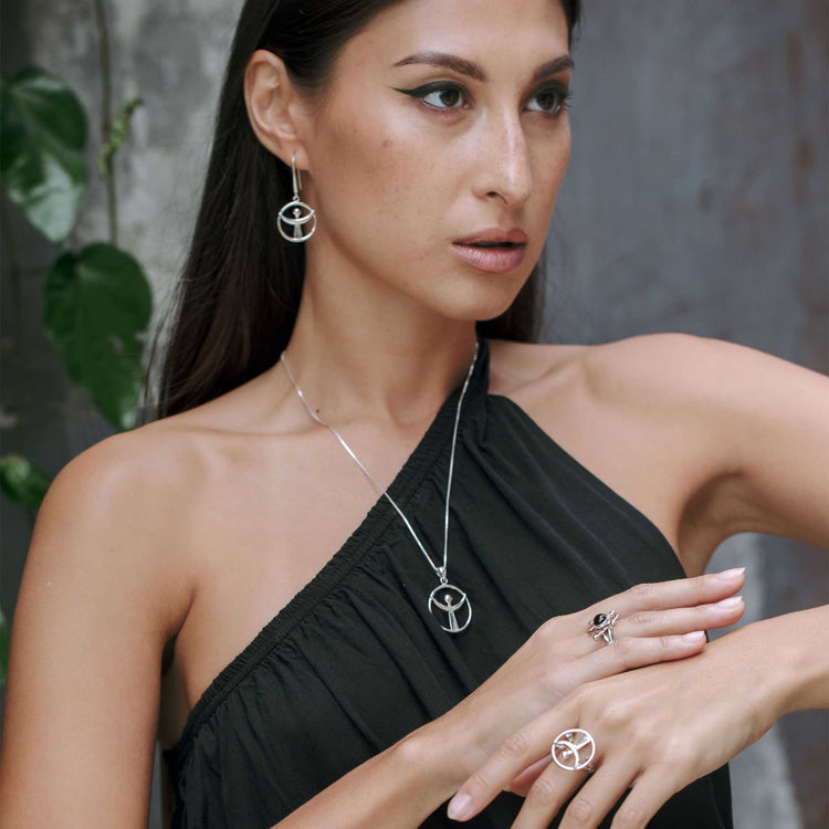 A woman in a one-shoulder black dress models Kaora Sandara Jewelry’s Releasing From the Deepest Fears Ring in Sterling Silver (Size S), with matching jewelry, set against a gray textured wall and green leaves.