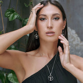 A woman with long dark hair wearing a one-shoulder black top poses before a leafy wall, her Kaora Sandara Jewelry "Releasing From the Deepest Fears" Sterling Silver 925 Pendant (Size S) and matching pieces gleaming in the light.