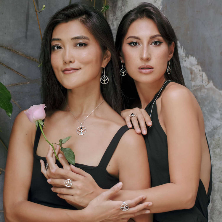 Two women in black pose by a rustic wall, one holding a pink rose. Both wear Kaora Sandara Jewelry's Releasing From the Deepest Fears Pendant in Sterling Silver with Black Agate, Size L, radiating calm and confidence.