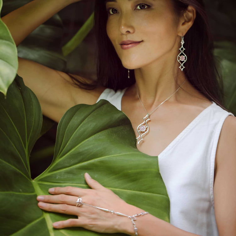 A woman in a white sleeveless top stands among lush green leaves, smiling softly as she gently touches a leaf. She wears Kaora Sandara Jewelry’s Quantum Shift NEW Ring – Sterling Silver with Rose Quartz, Size L.