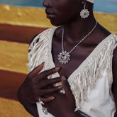 A woman in a fringed white top highlights Kaora Sandara Jewelry's Purity Earhooks – Sterling Silver with Crystal, Size S, along with a necklace and ring, set against a vibrant yellow and blue background. Her hands rest near her chest and necklace.