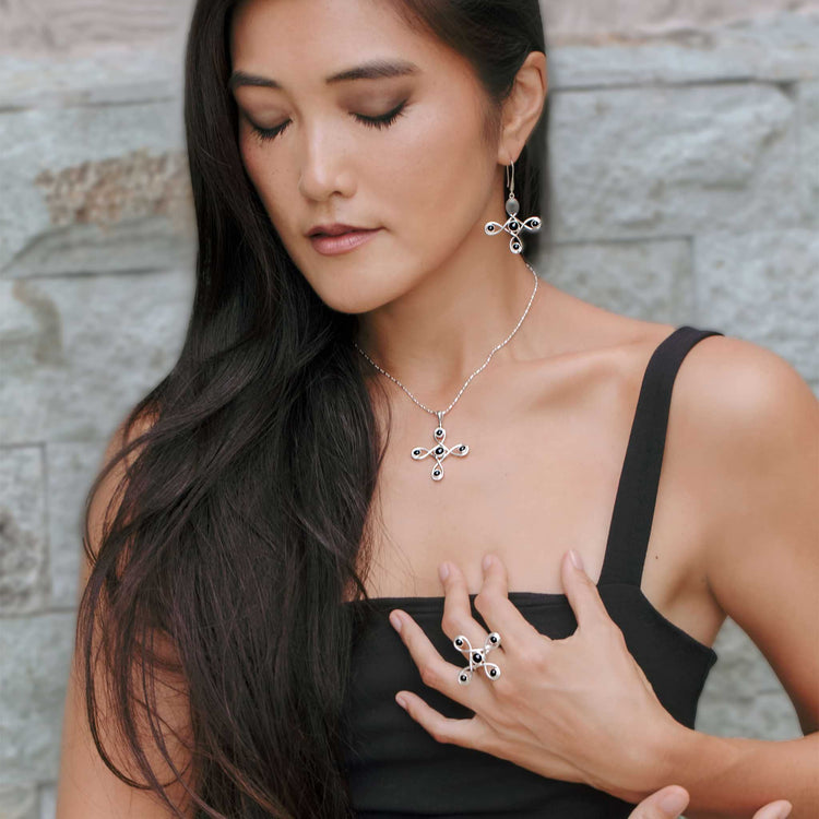 A woman with long dark hair and closed eyes poses against a stone wall in a black sleeveless top, wearing Kaora Sandara Jewelry: the Programs Clearing Pendant in Sterling Silver with Black Agate, plus matching earrings and ring.