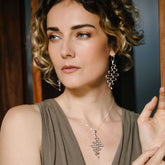 A woman with short, curly hair wears a sleeveless taupe dress and Kaora Sandara Jewelry’s Silver Earhooks Team Work earrings, gazing thoughtfully to the side with one hand resting on a wooden surface—ready for her team meeting.