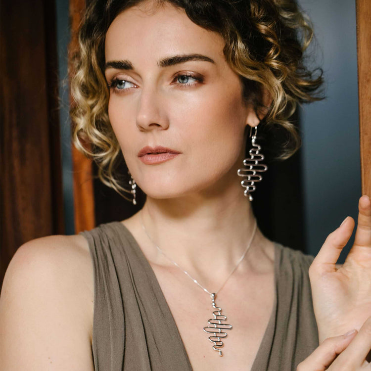 A woman with short, curly hair wears a sleeveless taupe dress and Kaora Sandara Jewelry’s Silver Earhooks Team Work earrings, gazing thoughtfully to the side with one hand resting on a wooden surface—ready for her team meeting.