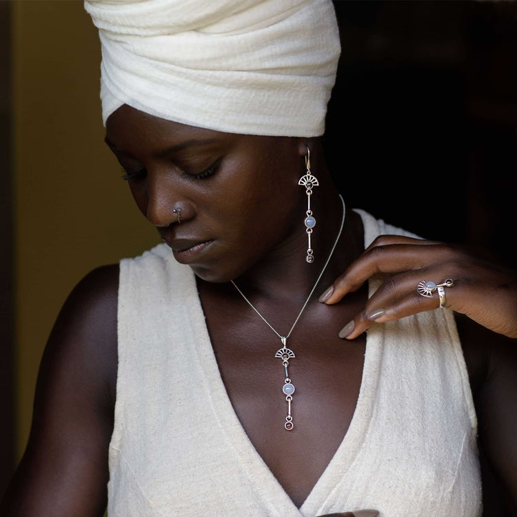 A woman in a white headwrap and sleeveless top looks down, highlighting Kaora Sandara Jewelry’s Peace of Mind Earhooks—Sterling Silver with Crystal & Chalcedony—that feature elegant circular designs.