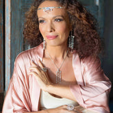A woman with curly hair wears a pink satin robe and silver jewelry, including the Magic Path Headband from Kaora Sandara Jewelry, adorned with crystals. She gazes thoughtfully to the side against a rustic wooden background.