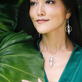 A woman in a green top stands by a large leaf, smiling softly while wearing Kaora Sandara Jewelry’s Intuition Pendant—Sterling Silver & Gold-Plated with Amethyst—and circular earrings.