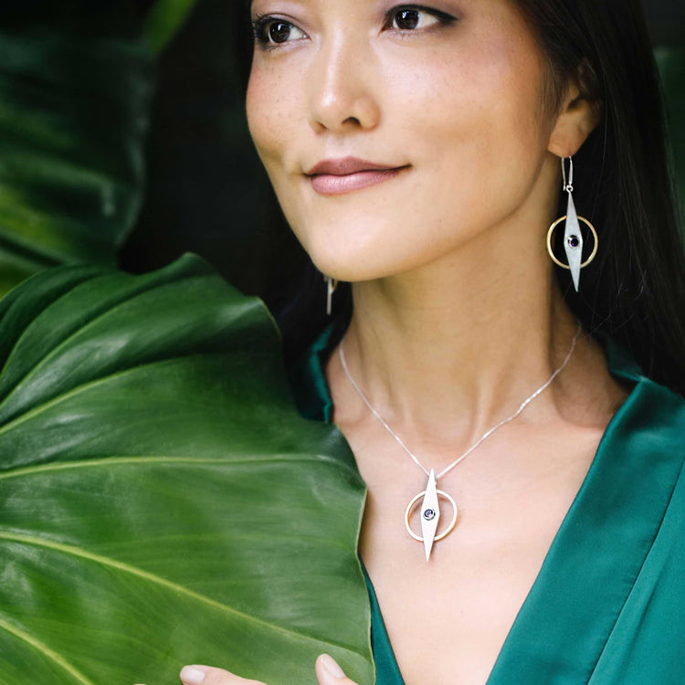 A woman in a green top stands by a large leaf, smiling softly while wearing Kaora Sandara Jewelry’s Intuition Pendant—Sterling Silver & Gold-Plated with Amethyst—and circular earrings.