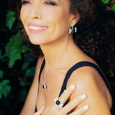 A woman with curly hair smiles, wearing a black top and the Kaora Sandara Jewelry Health Formula Ring—crafted from sterling silver with black agate and crystal—paired with matching jewelry, set against a backdrop of blurred greenery.