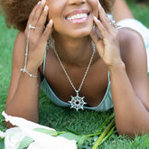 A woman smiles while lying on grass beside white lilies, wearing Kaora Sandara Jewelry's Freedom from the Ego Pendant in sterling silver with crystal, rose quartz, and peridot, along with rings and a bracelet.