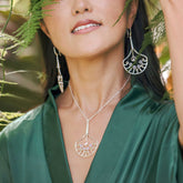 Amid lush greenery, a woman in a green satin dress models the Kaora Sandara Jewelry Body Relaxation Pendant—sterling silver with crystal, chalcedony & peridot—paired with bold silver earrings. Her lips are parted, face partially hidden by leaves.