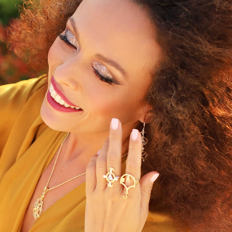 A woman with curly hair smiles, wearing shimmery eye makeup, pink lipstick, gold jewelry including Kaora Sandara Jewelry’s Abundance Flow Ring in sterling silver & gold-plated with crystal & citrine, plus a necklace and earrings, and a mustard yellow top.