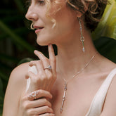 A woman with short curly hair models Kaora Sandara Jewelry’s State Ring—sterling silver with crystal & chalcedony—alongside silver drop earrings and a pendant necklace, set against lush green leaves.