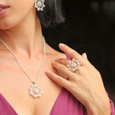 A woman wearing Kaora Sandara Jewelry’s Source of Love Ring—Sterling Silver with Crystal & Rose Quartz—alongside a matching flower pendant necklace and earrings, poses with her hand near her collarbone in a mauve V-neck top.