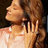 A woman with long wavy hair in a pink satin top smiles with closed eyes, wearing Kaora Sandara Jewelry’s Anima Earhooks—Sterling Silver with Crystal & Rose Quartz. Sunlight highlights her face and the elegant earrings.