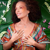 A smiling woman with curly hair wears vibrant clothing and the Anima Pendant – Sterling Silver with Crystal & Rose Quartz by Kaora Sandara Jewelry, lying on green leaves with white and purple flowers, hands gently resting on her chest.