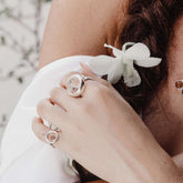 A woman's hand wears the Kaora Sandara Jewelry Silver Ring Crystal Peace with Crystal (Size L) as she holds a white orchid near her wavy brown hair, dressed in a white off-shoulder garment.