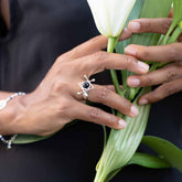 A person wears the Kaora Sandara Jewelry Energy Protection Ring—Sterling Silver with Black Agate—while holding a white lily, showcasing manicured hands and a dark sleeveless outfit for an elegant, protective style.