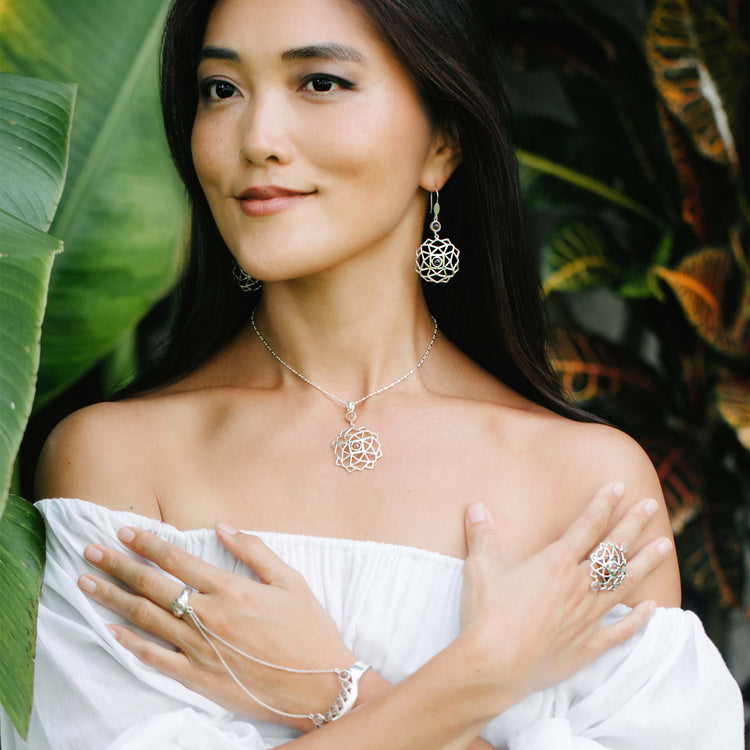 A woman in an off-shoulder white top poses among lush leaves, wearing Kaora Sandara Jewelry’s Sterling Silver Expansion Pendant with Crystal and matching geometric jewelry from the elegant Expansion series.