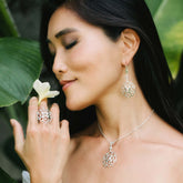 A woman with long dark hair stands by green leaves, peacefully holding a white flower. She wears Kaora Sandara Jewelry's Expansion Earhooks—sterling silver with crystal spheres—for a bold, geometric touch.