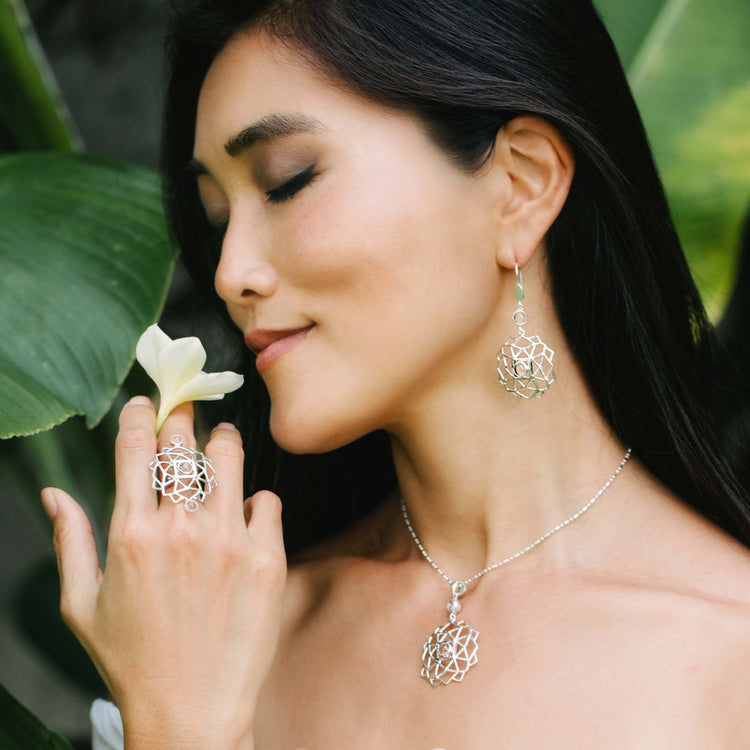 A woman with long dark hair stands by green leaves, peacefully holding a white flower. She wears Kaora Sandara Jewelry's Expansion Earhooks—sterling silver with crystal spheres—for a bold, geometric touch.