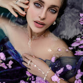 A woman with striking blue eyes lies partly submerged in water, wearing Kaora Sandara Jewelry's Silver Hand Chain Crystal Path Crystal Sphere Gemstones. Pink petals float around her, adding to the dreamy, ethereal mood.