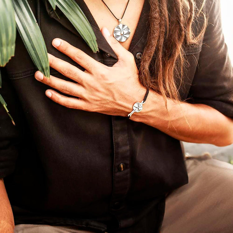 A person in a dark shirt rests their hand on their chest, displaying a round rock crystal pendant and a Kaora Sandara Jewelry Silver Male Bracelet On Dark Grey String with Code Abundance. Green leaves partly cover their shoulder.
