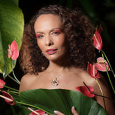 A woman with curly hair and bold makeup poses among pink anthuriums, holding a green leaf over her chest. She wears Kaora Sandara Jewelry’s Animus Pendant—sterling silver and gold-plated with pyrite—amid lush green foliage.
