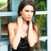 A woman with long brown hair in a black strapless dress poses by a window, wearing the Kaora Sandara Jewelry Silver Pendant Magic Star with Crystal Sphere - Size M, her hands touching her neck as she gazes thoughtfully into the distance.