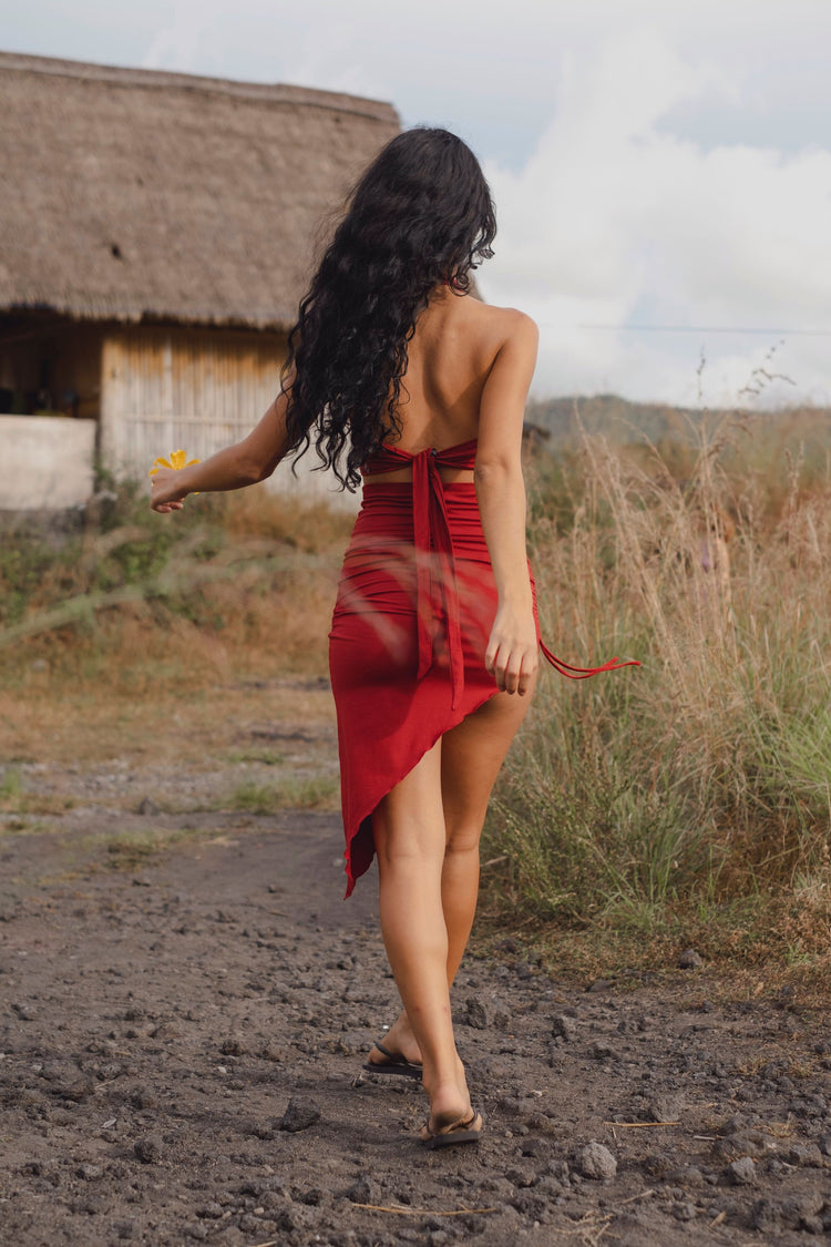 A woman with long, curly dark hair walks barefoot on a dirt path near tall grass and a rustic thatched-roof building, holding a yellow flower. She wears an Ikuti Red Venus Yoga Top—ring front halter crop in sustainable fabric. Photo is taken from behind.