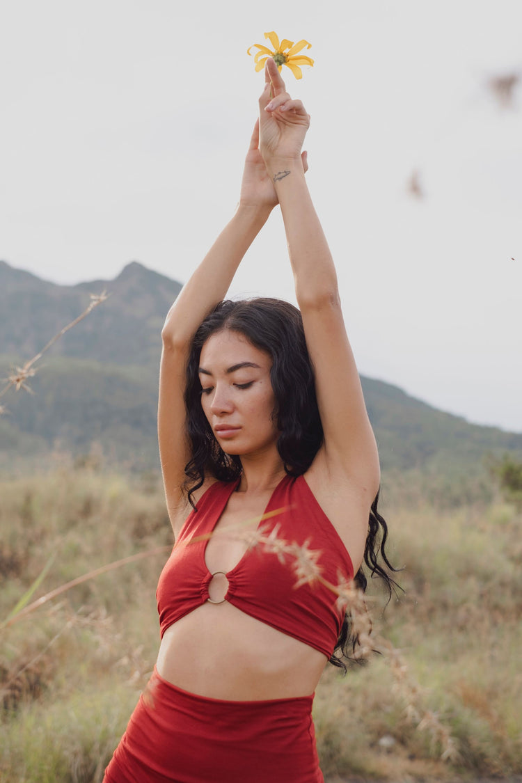 A woman with long dark hair and closed eyes stands outdoors, arms raised holding a yellow flower, wearing the Ikuti Red Venus Yoga Top—a retro, eco-friendly halter crop top—against a backdrop of mountains and grassy fields.