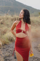 A woman outdoors on a dirt path wears the Ikuti Red Venus Yoga Top, a ring front halter crop top in eco-friendly fabric. She holds a yellow flower, looking relaxed, with grassy vegetation and mountains behind her.
