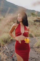 A woman stands outdoors on a rocky path, wearing the Ikuti Red Venus Yoga Top—a ring front halter crop—amid grassy fields and mountains. She holds a yellow flower and looks to the side, surrounded by tall dried grass.