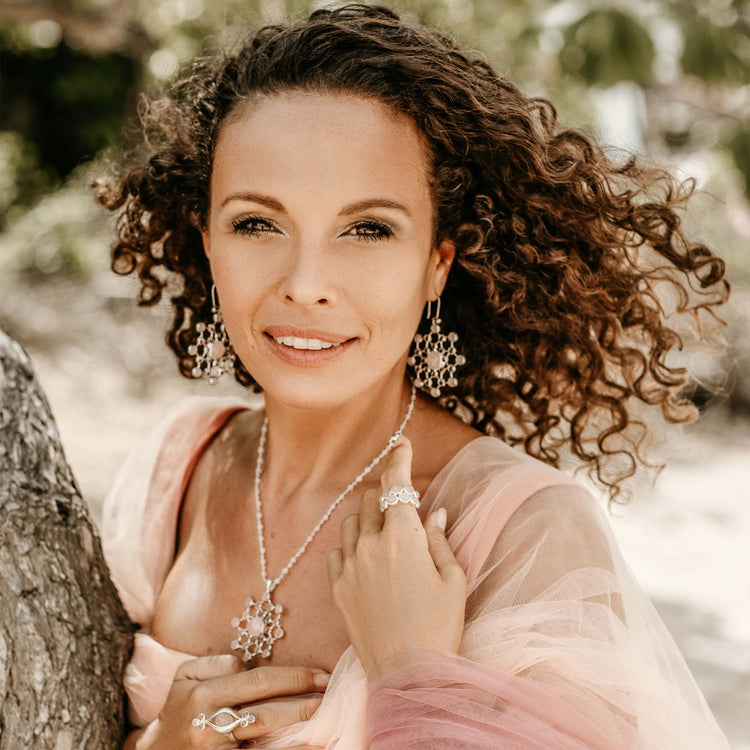 A woman with curly hair stands by a tree, wearing a sheer pink top and the Silver Ring The Dance with Rose Quartz by Kaora Sandara Jewelry. Sunlight filters through the background, highlighting her elegant, spiritual style.