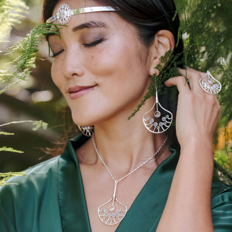 A woman with closed eyes wears the Kaora Sandara Jewelry Body Relaxation Ring—sterling silver with crystal, chalcedony, and peridot—smiling gently in a deep green top among vibrant foliage, radiating calm and relaxation.