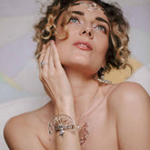 A woman with curly hair gazes upward, wearing the NEW! Higher Self Bracelet—Sterling Silver with Crystal & Chalcedony by Kaora Sandara Jewelry—on her wrist, evoking her higher self against a soft pastel background.