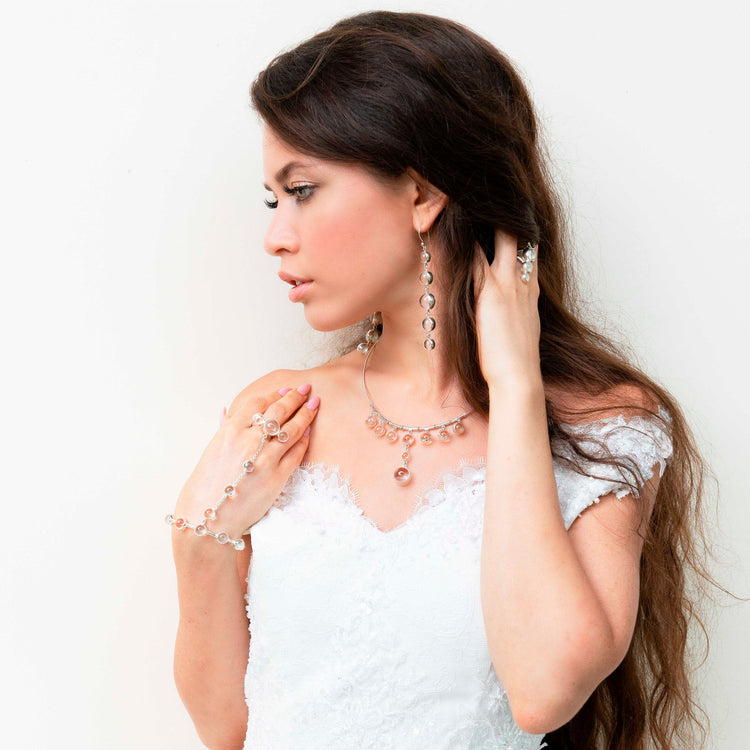 A woman in a white lace dress poses against a white background, modeling Kaora Sandara Jewelry’s Harmony Earhooks—sterling silver with crystal, size L. She has long brown hair and gazes to the side with one hand near her face.