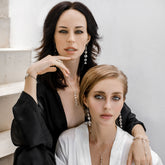 Two women sit closely on white stone steps in elegant black and white outfits, both wearing Kaora Sandara Jewelry's Harmony Earhooks—Sterling Silver with Crystal, Size S. They look directly at the camera.