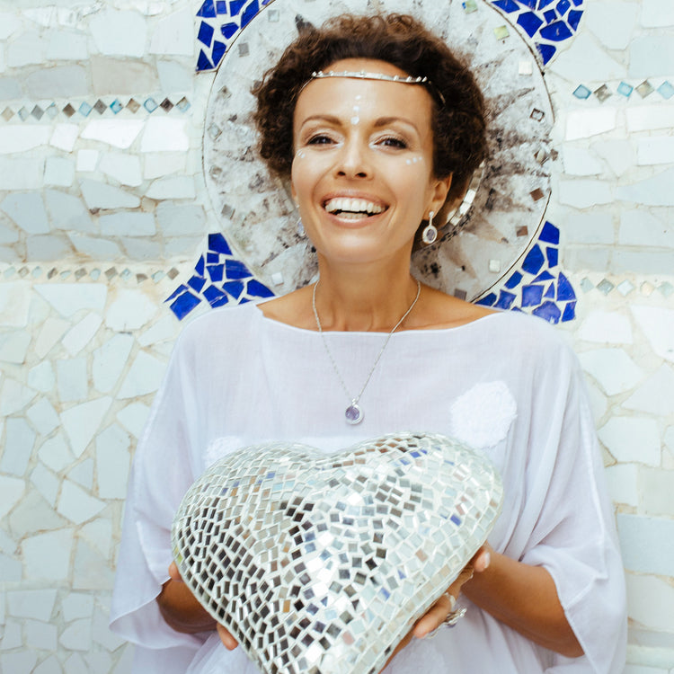 A smiling woman in a white dress wears the Kaora Sandara Jewelry Silver Pendant Crystal Peace with Silver-Chalcedony Chain and Chalcedony Sphere Gemstones (Size L), standing before a white tiled wall with blue accents.
