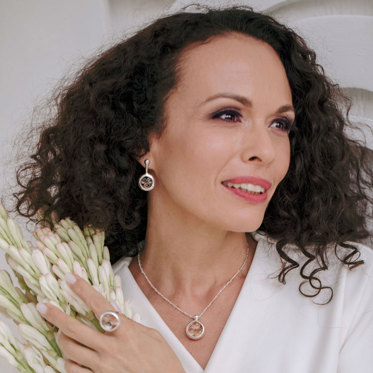 A woman with curly dark hair in white attire smiles serenely while holding white flowers, wearing Kaora Sandara Jewelry’s Silver Pendant Crystal Peace with Silver-Crystal Chain and Crystal Sphere Gemstones, size L.
