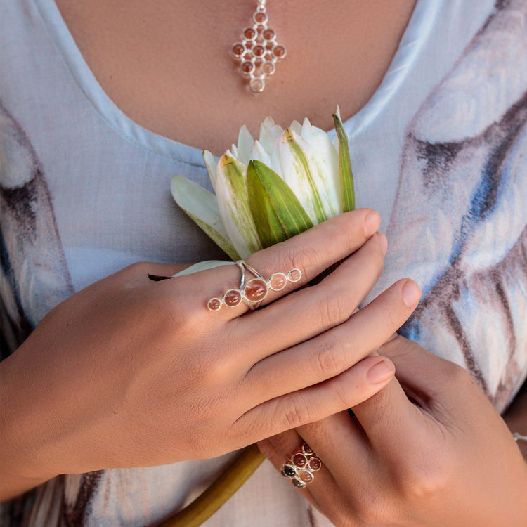 Adorned with a Kaora Sandara Jewelry Silver Ring Harmony with Crystal Sphere Gemstones (Size S) and a matching necklace, a person in a light patterned top holds a white flower close to their chest.