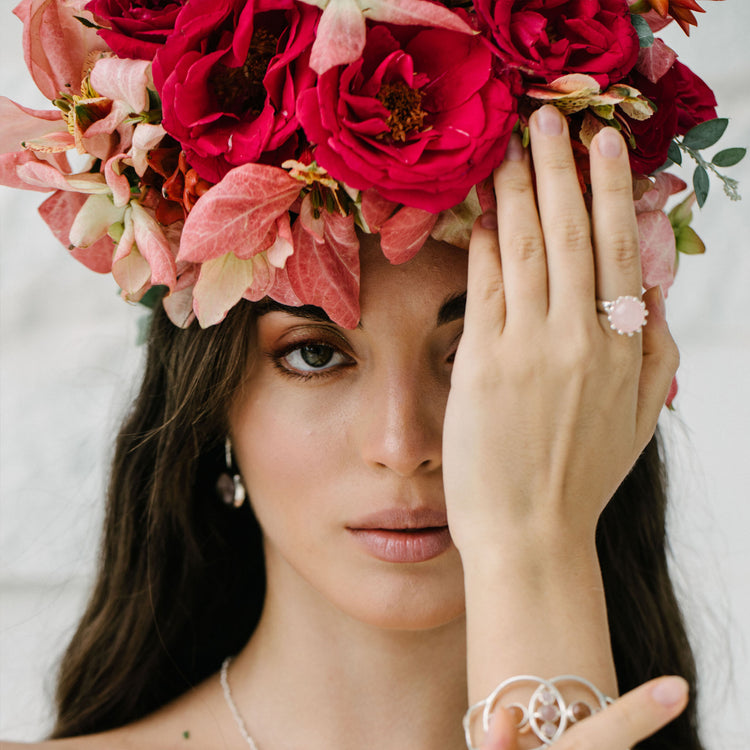 A woman with long dark hair wears a crown of red and pink flowers. Covering one eye, she shows off the Kaora Sandara Jewelry Silver Ring I'M A Queen with Rose Quartz (Size S), reflecting calm elegance and unconditional love.