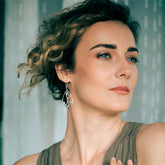 A woman with short, curly hair gazes thoughtfully, wearing a sleeveless taupe top and Kaora Sandara Jewelry’s Silver Earhooks Self-Worth – Crystal Gemstone. She stands before a softly lit sheer curtain backdrop.