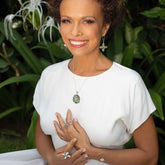 A curly-haired woman in a white dress smiles outdoors, wearing Kaora Sandara Jewelry's Power's Activation Earhooks—sterling silver with crystal—surrounded by green plants and white flowers.