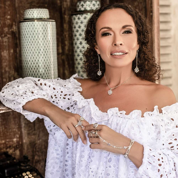 A woman with curly hair, wearing a white off-the-shoulder dress and the Kaora Sandara Jewelry Silver Hand Chain Goddess Path with Chalcedony & Crystal Sphere, stands indoors near vintage decor, smiling at the camera.