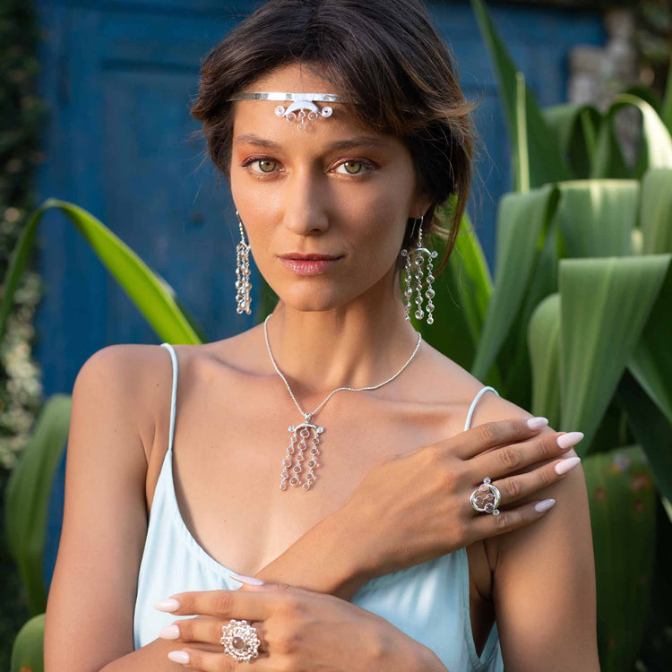 A woman in a light blue dress poses outdoors among large green leaves, wearing Kaora Sandara Jewelry’s Magic Path Earhooks—Sterling Silver 925 with Crystal Gemstones—along with a matching headpiece, pendant necklace, and statement rings.