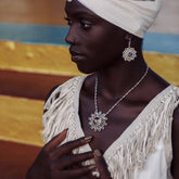 A woman in a white head wrap and fringed outfit models Kaora Sandara Jewelry’s I am in Everything Pendant—sterling silver with crystal—alongside matching earrings, against a vibrant striped background.