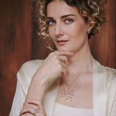 A woman with curly hair in a cream top poses with her hand near her face, showcasing Kaora Sandara Jewelry's Perfection of Life Pendant—Sterling Silver with Crystal & Chalcedony—plus matching earrings and rings, against a wooden backdrop.