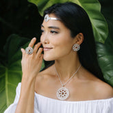 A woman in an off-shoulder white top wears Kaora Sandara Jewelry's Purity Ring—Sterling Silver with Crystal, Size L—and matching jewelry with circular designs, brown accents, and rhinestones. She poses before lush green leaves.