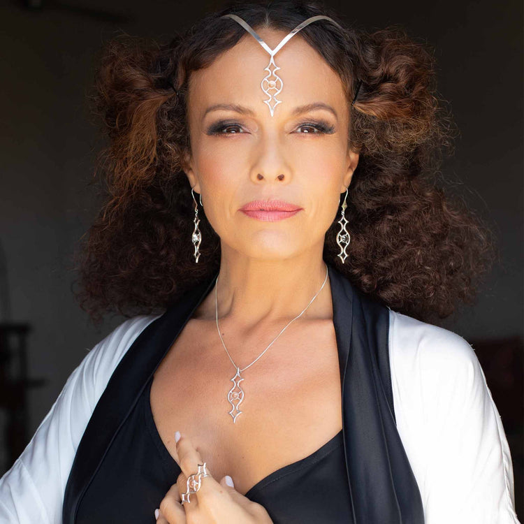 A woman with curly hair wears the Quantum Shift NEW Pendant by Kaora Sandara Jewelry, crafted in sterling silver with crystal gemstones. She pairs it with a black top and white shawl, posing confidently against a dark background.